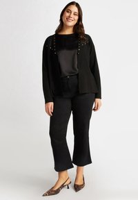 Schwarze, offene Strickjacke mit goldenen Nieten, darüber ein schwarzes Satinoberteil. Schwarze, weit geschnittene Hose und spitze, leopardengemusterte Slingback-Schuhe.