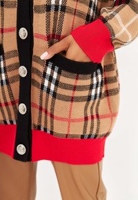 Cárdigan de cuadros beige y negro con puños y dobladillo de canalé rojos, que presenta grandes botones plateados y bolsillos frontales. Textura de punto suave.