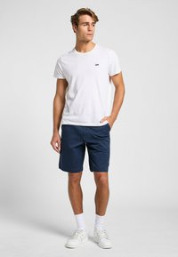 Hvid bomulds T-shirt med et lille sort "Lee" logo, parret med marineblå shorts. Modellen bærer hvide sneakers og ankelsokker.