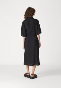 Black, knee-length dress with a relaxed fit, short sleeves, and button-down collar; made of lightweight, textured fabric. Paired with black sandals.