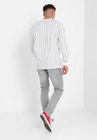 Camicia bianca a maniche lunghe con righe verticali nere, abbinata a pantaloni grigi e scarpe rosse. L'immagine mostra il retro dell'outfit.