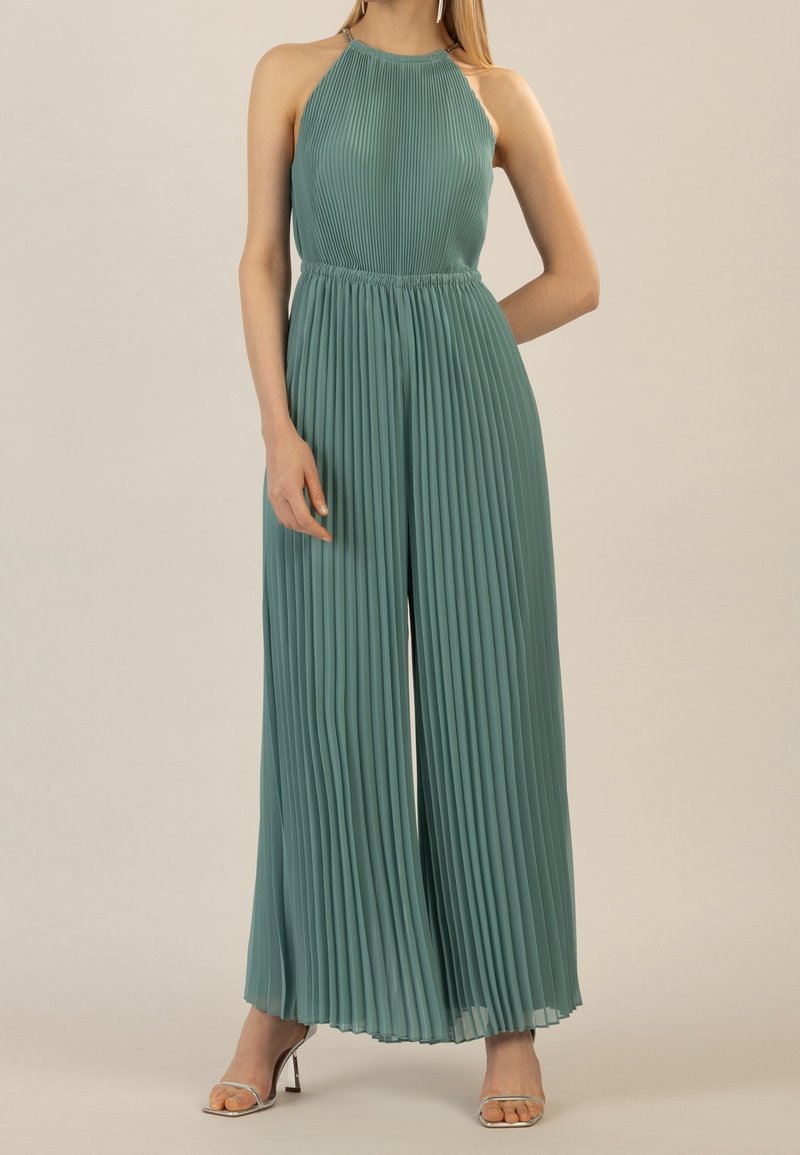 Femme portant une combinaison sans manches plissée bleu sarcelle à col halter et des talons ouverts argentés, debout devant un fond beige uni.