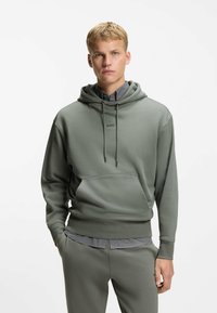Hooded sweatshirt in muted green fabric, featuring a front pocket, drawstrings, and subtle logo. Worn with matching pants showcasing a textured waistband.