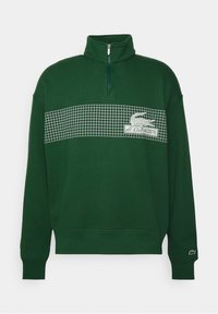 Sweat-shirt vert avec un col haut, une demi-fermeture éclair et un motif en grille sur la poitrine, présentant un logo crocodile brodé sur la manche gauche.