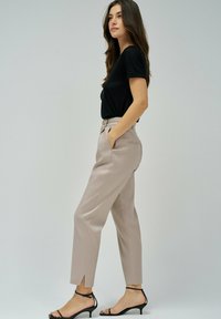 Mujer con una blusa negra de manga corta, pantalones beige de tiro alto y ajustados con bolsillos laterales, y sandalias negras de tacón bajo, de pie de lado.