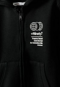 Černá mikina na zip vyrobená z měkké látky. Má bílou grafiku s designem zeměkoule a textem: „+Ninety7 Zlepši se, uč se nové věci, vydej se na divoká místa.“