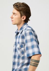 Lyhythihainen ruudullinen paita, jossa on sinisiä ja valkoisia sävyjä. Eteisessä on kaksi nappikiinnityksellä varustettua taskua ja klassinen kauluskamari.