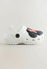 Zapato estilo zueco blanco con suela negra de lunares, correa ajustable en el talón y un lazo rojo y negro decorativo en la parte delantera.