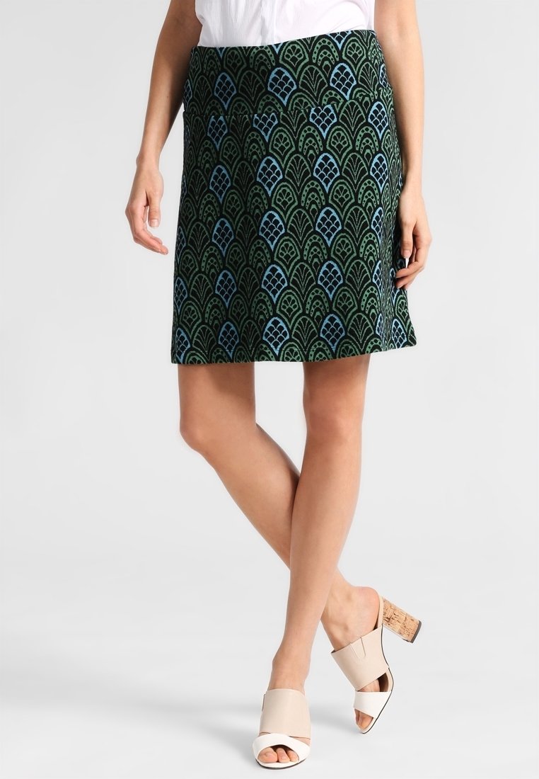 Femme portant une jupe vert foncé longueur genou avec des motifs de feuilles bleues et des sandales beiges à talons blocs et bouts ouverts, debout jambes croisées.