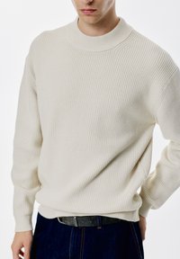 Cremfarbener Rippstrickpullover mit hohem Rundhalsausschnitt, schmalen Bündchen und lockerer Passform, kombiniert mit blauen Denimhosen und einem dunklen Gürtel.