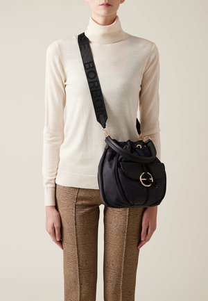 Borsa in tessuto nero texturizzato con una chiusura circolare dorata, abbinata a pantaloni marroni a fantasia e un maglione a collo alto color crema. Tracolla regolabile.