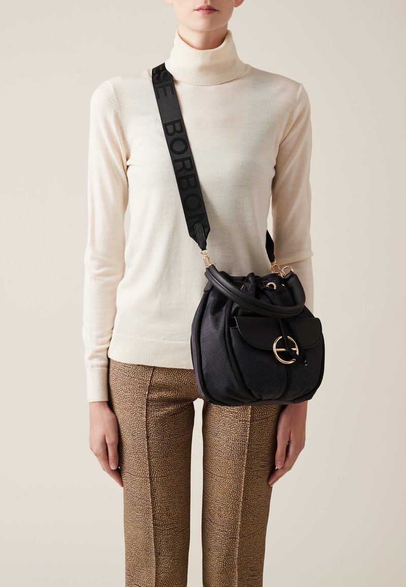 Tkaninowa torba w czarnym kolorze o teksturowanej powierzchni, z okrągłym zapięciem w kolorze złotym, zestawiona ze wzorzystymi brązowymi spodniami i kremowym swetrem z golfem. Regulowany pasek.