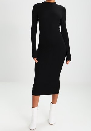 Robe midi noire côtelée avec manches longues et col montant, présentant une silhouette ajustée et des poignets de manches allongés. Portée avec des bottines blanches à la cheville.