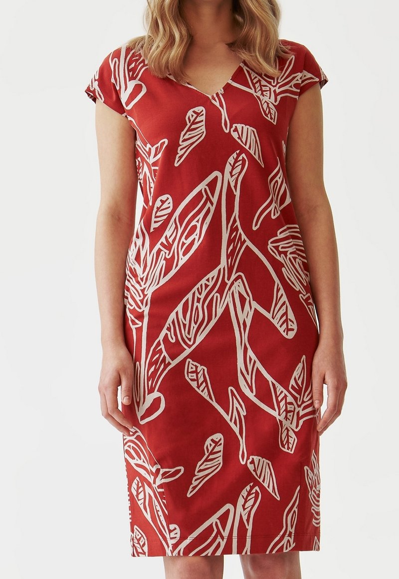Femme portant une robe rouge mi-longue à manches courtes avec des motifs abstraits blancs de feuilles et de branches sur un fond uni.