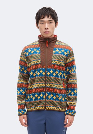 Mann trägt eine bunt gemusterte Zip-Up-Fleecejacke mit braunem Brustpaneel und hohem Kragen, steht vor einem schlichten hellen Hintergrund.