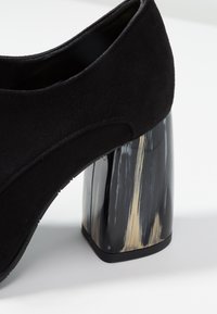 Svart mocka klackskor med en framträdande transparent blockklack som har svarta och gyllene streck. Slät textur och rund tådesign.