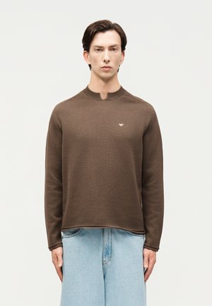 Άνδρας που φοράει καφέ πουλόβερ με μακριά μανίκια και μικρό λογότυπο στο στήθος και ανοιχτό μπλε τζιν παντελόνι με φαρδιά μπατζάκια, στέκεται μπροστά σε απλό φόντο.