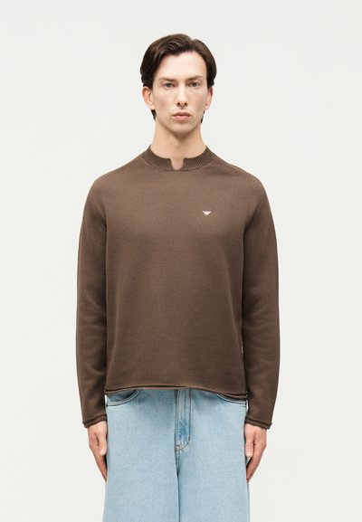 Homme portant un pull marron à manches longues avec un petit logo sur la poitrine et un jean large bleu clair, debout devant un fond uni.