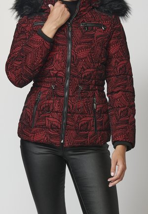 Femme portant une doudoune rouge et noire à motif avec une capuche bordée de fourrure et un pantalon noir aspect cuir, main près du col.