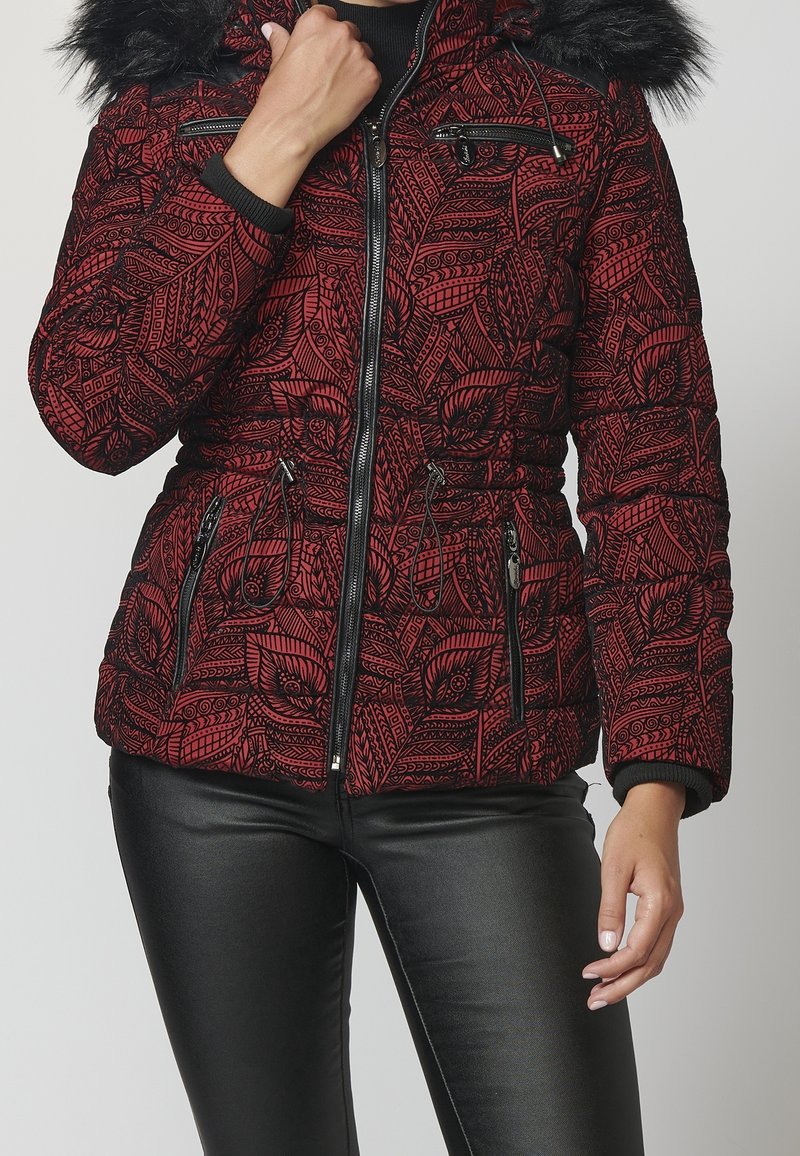 Femme portant une doudoune rouge et noire à motif avec une capuche bordée de fourrure et un pantalon noir aspect cuir, main près du col.
