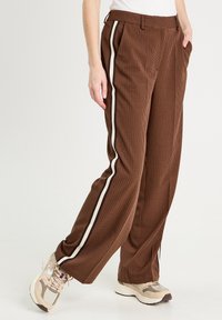 Pantaloni a quadri marroni con una vestibilità comoda, caratterizzati da strisce laterali bianche e nere, pieghe e tasche laterali. Indossati con sneakers beige.