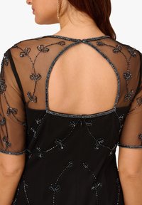 Schwarzes Kleid mit transparenten, bestickten Ärmeln und zarten, floral gestalteten Perlendetails. Der V-förmige Rücken mit einem Knopfverschluss verleiht Einzigartigkeit.
