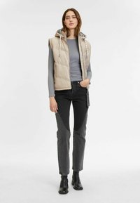 Beige gepolsterte Weste mit Kapuze, graues geripptes Oberteil und schwarze Jeans mit strukturiertem Muster. Schwarze Stiefeletten runden das Outfit ab.