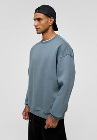 Lichtblauwe oversized sweatshirt met een ronde halslijn en verlaagde schouders, gemaakt van zacht materiaal. Draagt samen met zwarte broek, met een geprofileerd ontwerp.