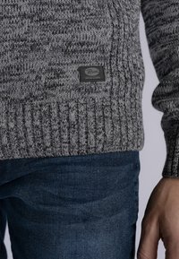 Maglione lavorato a maglia grigio con un motivo testurizzato. Orlo e polsini a coste. Etichetta nera del marchio cucita sul davanti. Abbinato a jeans blu scuro.