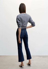 Suéter gris corto, jeans de mezclilla oscuros de cintura alta con dobladillo acampanado y tacones de color claro. Diseño simple con una textura sutil.