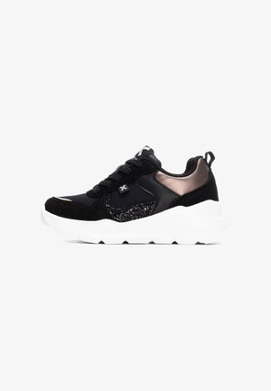 Zapatilla deportiva negra que presenta una combinación de materiales lisos y texturizados, una suela blanca y sutiles acentos metálicos en el talón.