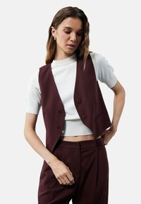 Gilet ajusté bordeaux à deux boutons, porté par-dessus un haut en tricot blanc. Texture lisse et design structuré avec une coupe courte.