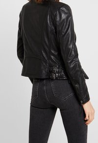 Veste en cuir noir au design ajusté, avec des boucles latérales et des poignets zippés, finition texturée et détails de couture.