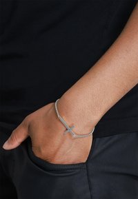 Sølvkjede armbånd med en horisontal korsanheng, vist på en hånd mot en svart skjorte. Glatt tekstur og minimalistisk design.
