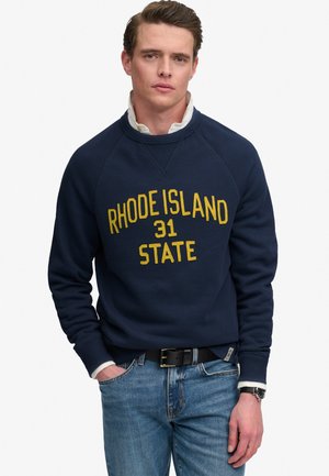 Młody mężczyzna w granatowej bluzie z napisem „Rhode Island 31 State” w żółtym kolorze, białej koszuli z kołnierzykiem, niebieskich dżinsach, czarnym pasku i zegarku na nadgarstku.