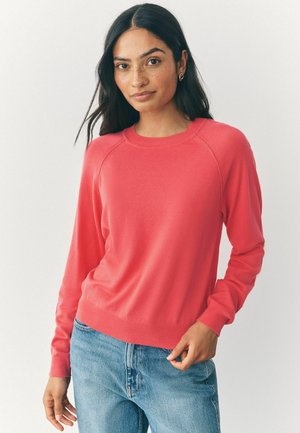 Korallenfarbener Strickpullover mit Rundhalsausschnitt und Raglanärmeln. Weiche Textur, tailliert, kombiniert mit blauen Jeans. Einfaches Design.