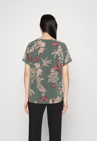 Mujer vista desde atrás con una blusa verde de manga corta con estampado de flores y mariposas rosas y pantalones negros, contra un fondo liso.
