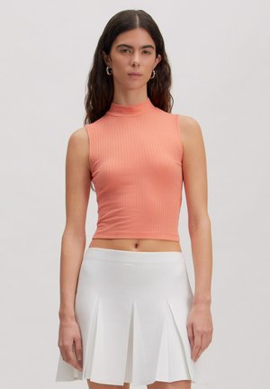 Jonge vrouw met lang donker haar, gekleed in een koraalrode mouwloze geribbelde crop top en een witte geplooide rok, staand tegen een effen achtergrond.