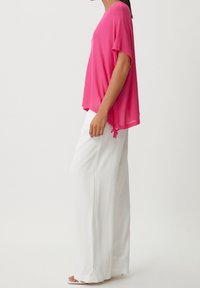 Top rosa a maniche corte con vestibilità ampia e dettaglio di nodo laterale, abbinato a pantaloni bianchi a gamba larga, caratterizzati da una texture liscia e un design informale.
