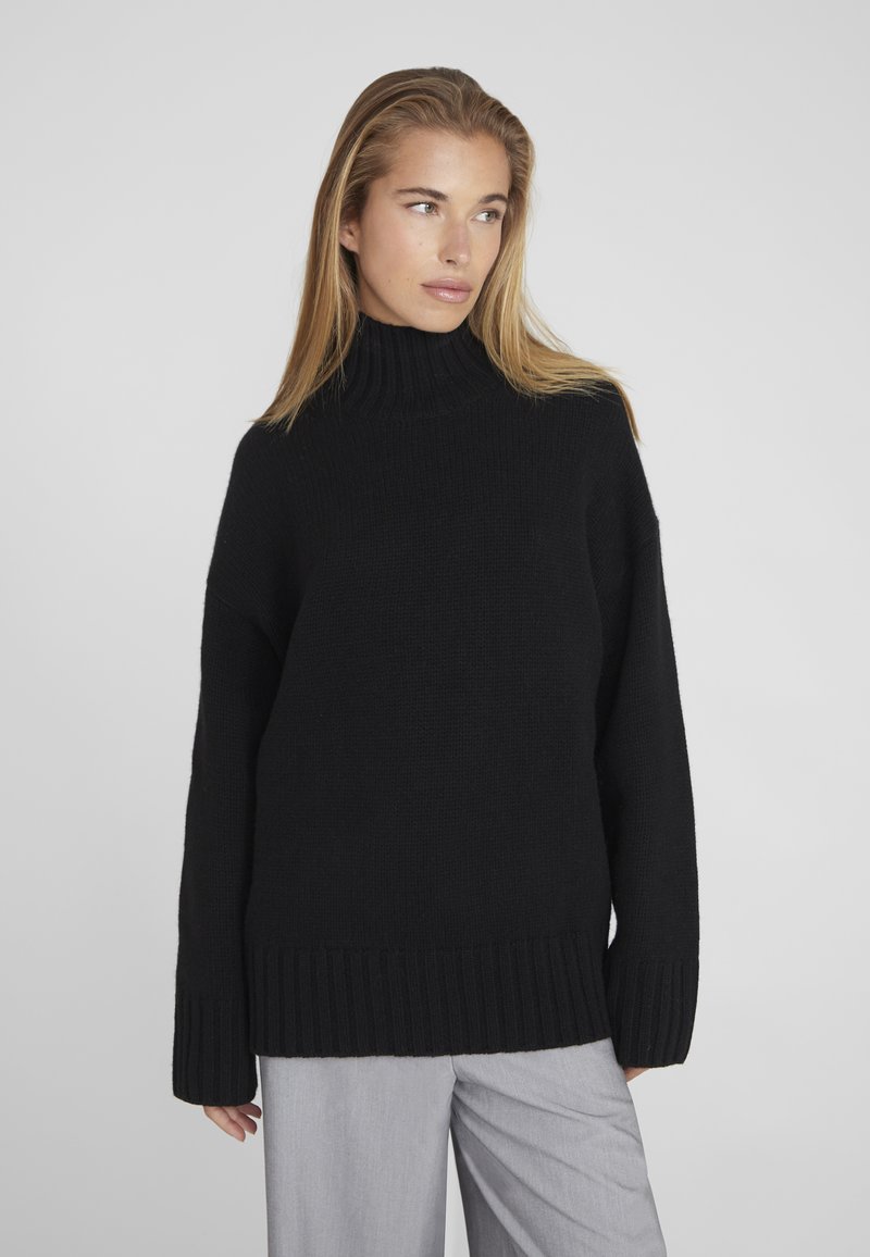 Pull en maille noir avec un col côtelé haut, coupe ample et poignets côtelés. Associé à un pantalon léger gris, avec une texture lisse.