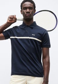 Μπλε ναυτικό πόλο με λευκή οριζόντια ρίγα και λογότυπο "BOSS"; κοντά μανίκια, γιακάς, συνοδευόμενο από σορτς κρεμ χρώματος, κρατώντας ρακέτα του τένις.