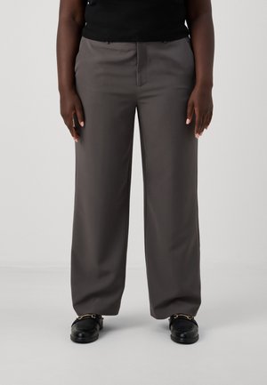 Graue weite Hosen aus einem glatten Stoff, mit einem Reißverschluss vorne, Gürtelschlaufen und einer taillierten Passform. Getragen mit schwarzen Schuhen.