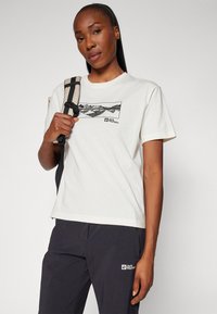 Crèmekleurig katoenen t-shirt met een grafisch ontwerp van een berglandschap. Het model houdt een rugzak vast en draagt donkere outdoor broeken met branding.