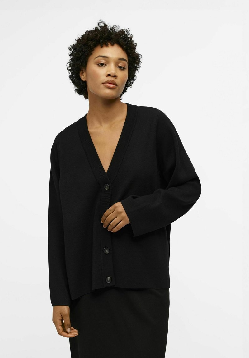 Cardigan noir avec un col en V, coupe ample, manches longues et quatre boutons à l'avant. Fabriqué en matériau texturé, associé à une jupe noire.