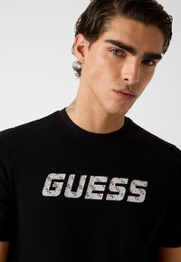 Zwart T-shirt met korte mouwen en een groot, wit, patroon met het "GUESS"-logo op de voorkant, gemaakt van zacht katoenen stof.