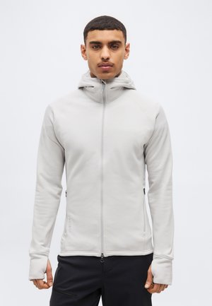 Bărbat purtând o jachetă ușoară gri deschis cu fermoar, glugă și mâneci cu orificii pentru degete, stând în fața unui fundal simplu alb.