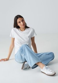 Witte katoenen t-shirt, relaxte pasvorm, gecombineerd met lichtblauwe jeans en witte sneakers met grijze accenten, zittend op een grijze vloer.