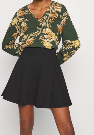Langärmlige grüne Bluse mit gelbem Blumenmuster, kombiniert mit einem schwarzen ausgestellten Rock. Die Bluse hat ein V-Ausschnitt-Design und eine lockere Passform.