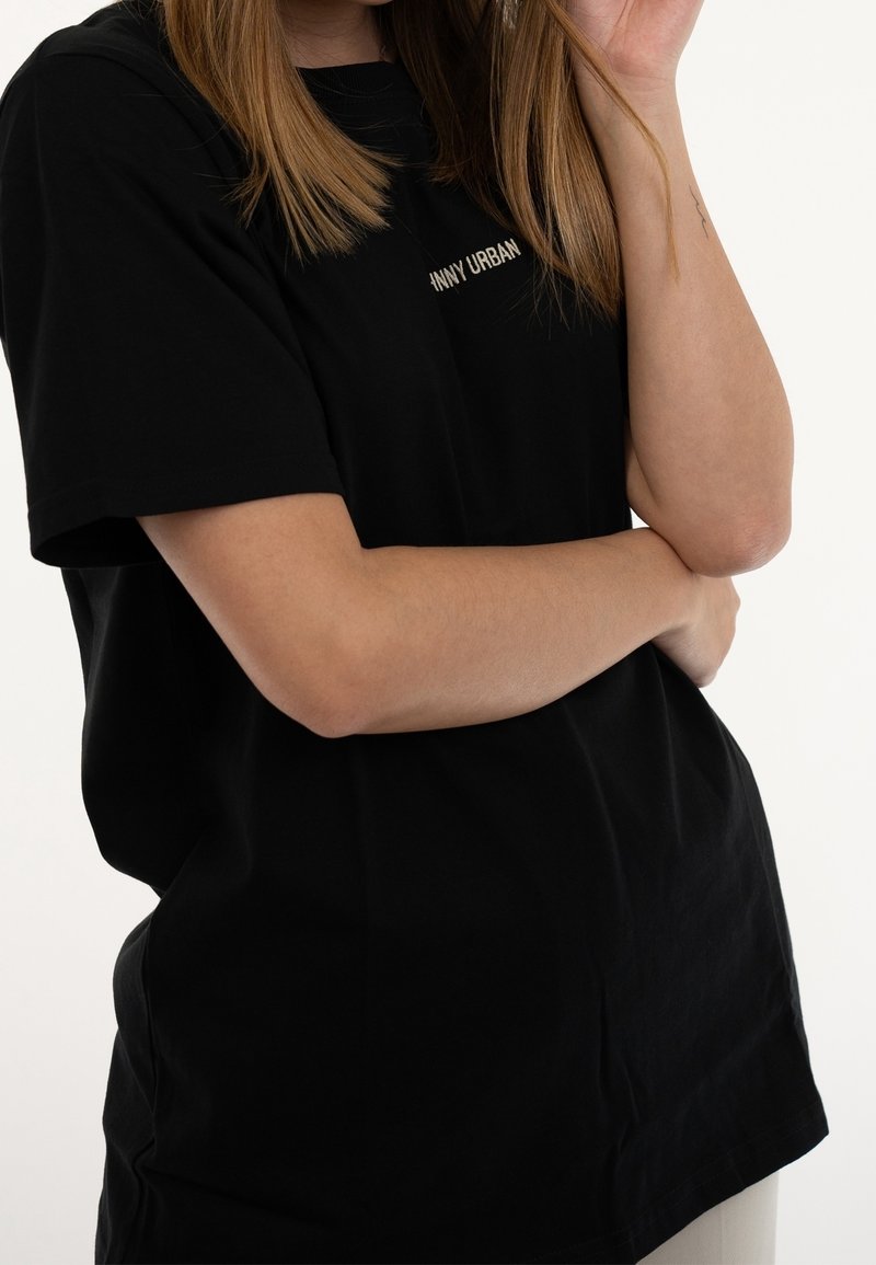 Personne portant un t-shirt noir "Vanny Urban" avec les bras croisés, cheveux châtain clair et un petit tatouage sur l'avant-bras gauche.
