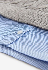 Chemise boutonnée à rayures bleues et blanches pliée, partiellement recouverte par un pull en tricot beige plié.